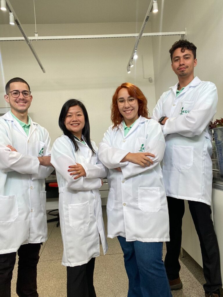 Grupo de quatro jovens cientistas, duas mulheres e dois homens, vestindo jalecos brancos, em um laboratório. 