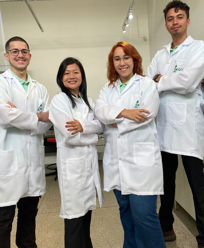 Grupo de quatro jovens cientistas, duas mulheres e dois homens, vestindo jalecos brancos, em um laboratório.